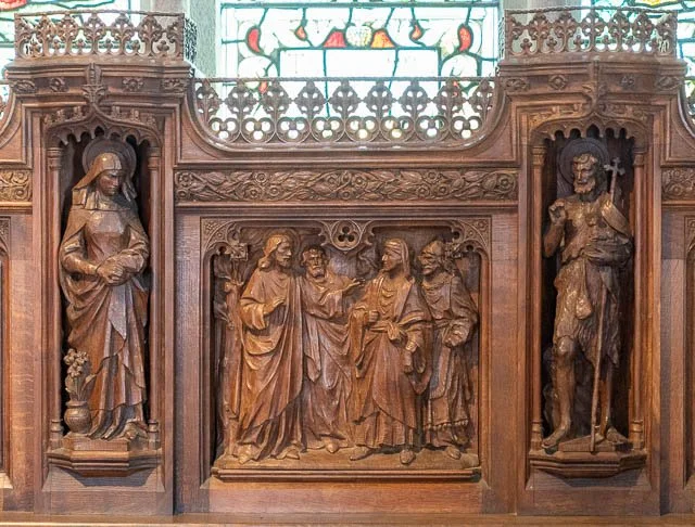 Wooden church altar featuring carved religious scenes, with figures of saints and angels, and stained glass window in the background.