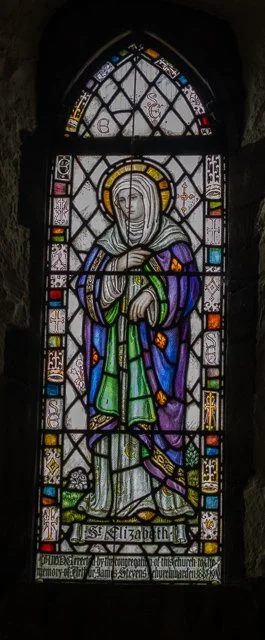 Stained glass window depicting a female saint in a colorful robe with a halo, holding a cross, with decorative geometric patterns and biblical symbols around her.
