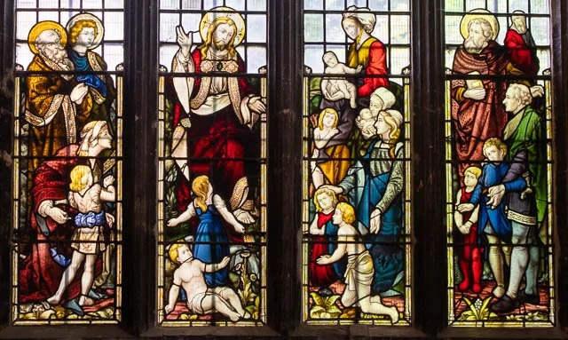 Colorful stained glass window depicting a biblical scene with Jesus surrounded by children and adults.