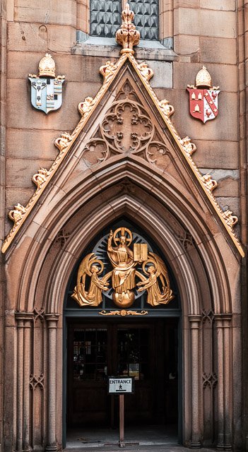Gothic archway entrance to a building with a gold religious sculpture of three figures inside the arch, flanked by two shields on the stone facade.