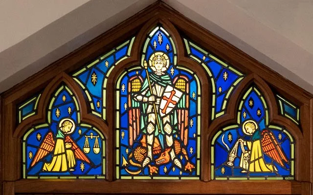 Colorful stained glass window depicting a saint in armor with a sword, flanked by angels holding scales and a sword.