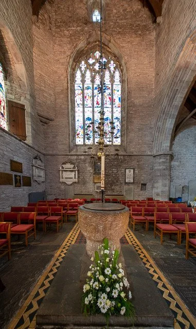 Inside a church with a stone baptismal font at the center surrounded by red chairs, a large stained glass window in the background, and a hanging cherub-shaped lamp. Flowers are placed at the base of the font.