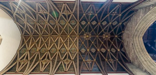 Looking up at a gothic cathedral ceiling with intricate wooden and stained glass designs.