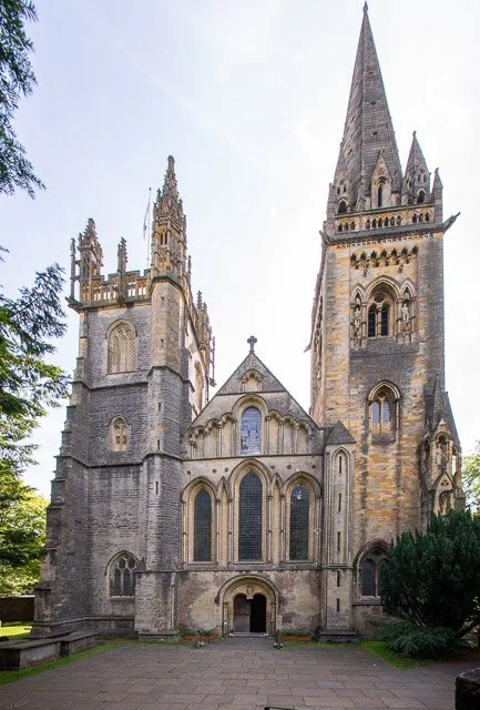 Photo of a large, historic Gothic-style cathedral with tall spires, arched windows, and ornate stone detailing, surrounded by trees.