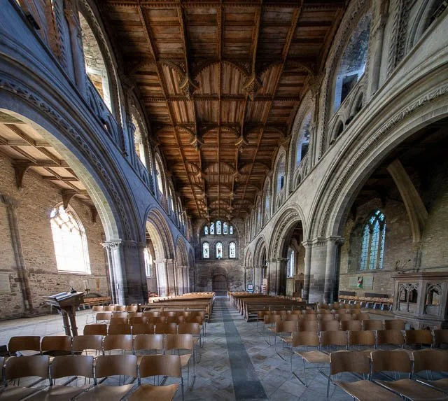 Interior of a historic church or cathedral with rows of chairs, arched windows, and detailed stone walls, wooden ceiling.