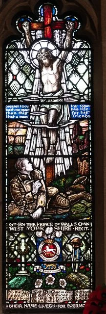 Stained glass window depicting Jesus Christ on the cross, with a figure of a man praying at the bottom, and various decorative elements including a shield and a cross.