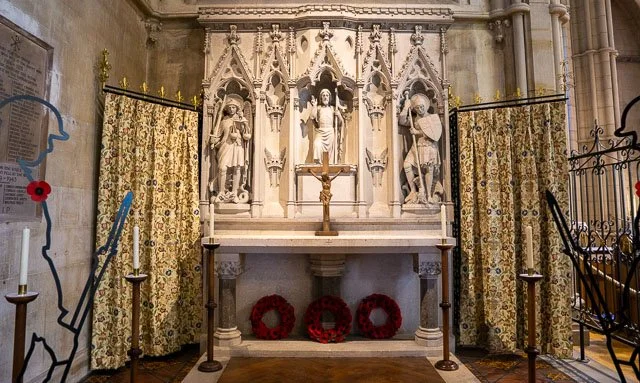 A church altar with a central crucifix, flanked by statues of saints, decorated with ornate curtains and candles on either side, with floral wreaths below.