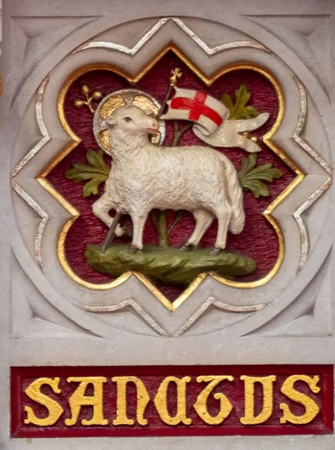Relief sculpture of a lamb with a halo and a flag, surrounded by decorative elements and the word 'SANDGS' in gold on a red background.