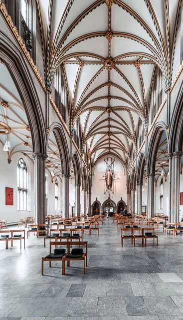 Interior of a grand Gothic-style cathedral with pointed arches, ribbed vaults, and tall stained-glass windows, arranged for a religious service with chairs and an altar at the front.