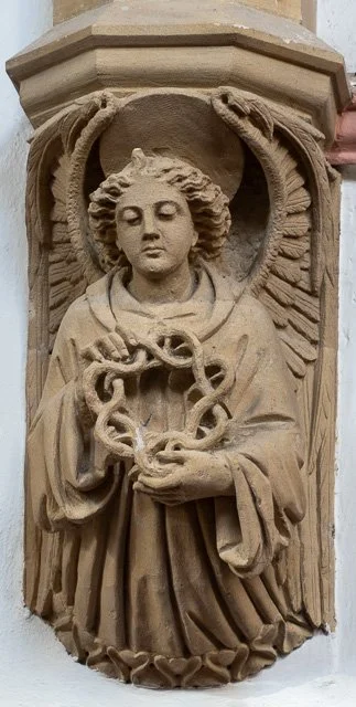 Stone relief sculpture of an angel with wings, holding a chain with a snake head, surrounded by a circular frame, mounted on a column.