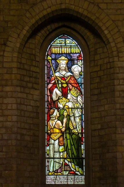 Colorful stained glass window depicting a crowned figure holding a scepter, surrounded by other figures, in a church setting.