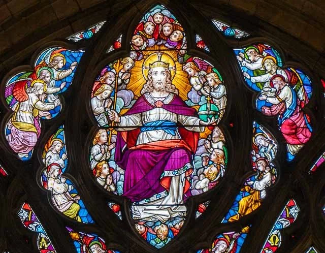 Stained glass window depicting Jesus Christ with a crown of rays, surrounded by angels and saints, in a Gothic style.