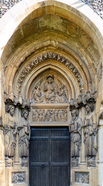 Stone religious sculpture depicting a central throne with seated figure flanked by two standing figures, surrounded by five standing figures, arched in Gothic style, above a black wooden door.