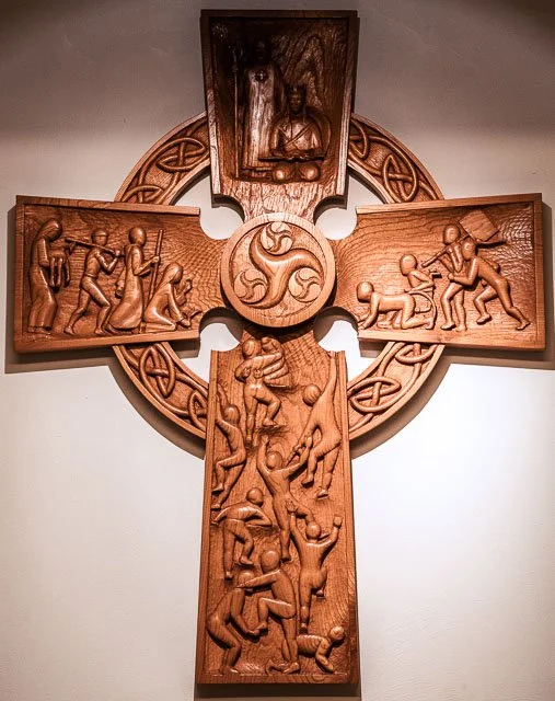 Wooden Celtic cross with carved scenes of people working, fighting, and celebrating, and a central triskelion symbol.
