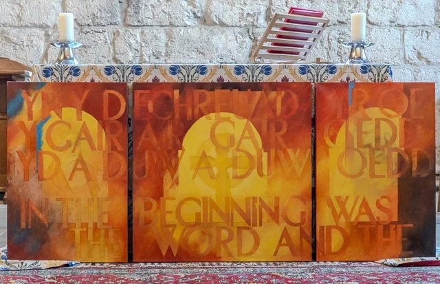 A three-paneled artwork with an orange and yellow abstract background, featuring overlaid text in varying shades saying, 'You can start again. The beginning was the word and the', with candles on each end on a table in front of a brick wall.