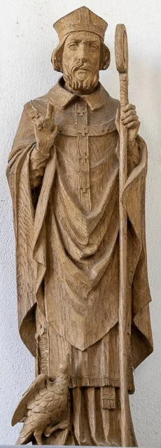 Wooden sculpture of a regal figure dressed in historical clothing, holding a staff and making a hand gesture, with a bird at his feet.