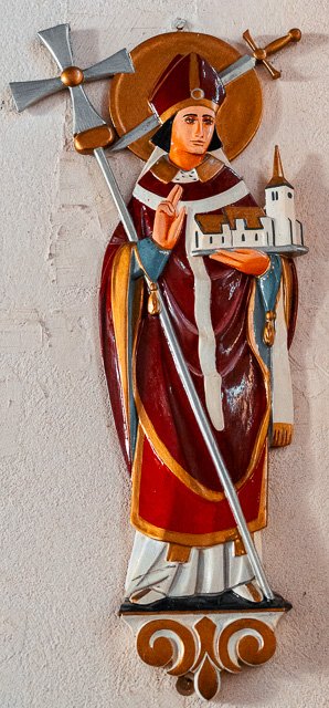 Colorful sculpture of a saint or religious figure holding a tower, dressed in traditional robes, with a halo and a windmill above the head, mounted on a wall.