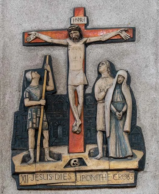 A wooden bas-relief depicting the crucifixion of Jesus Christ, with Jesus on the cross and two figures, a man and a woman, standing beside him.