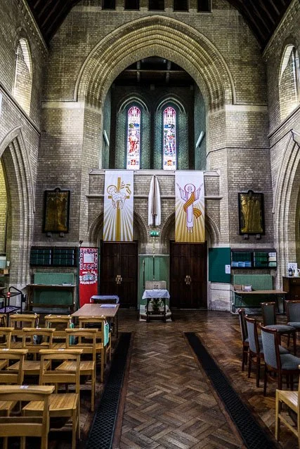 Interior view of a church with rows of wooden chairs, a small altar, and stained glass windows at the far end. The church has high brick walls and arched ceilings, with banners displaying religious symbols hanging above the entrance.