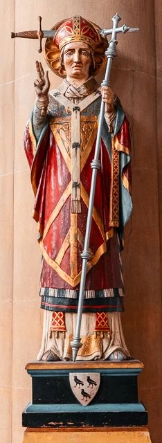 Statue of a saint holding a staff with a cross, dressed in long robes with intricate patterns, and making a gesture with the right hand, standing on a pedestal with a crest.