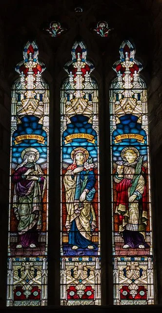 Colorful stained glass church window depicting three religious figures seated, with intricate designs and text banners above.