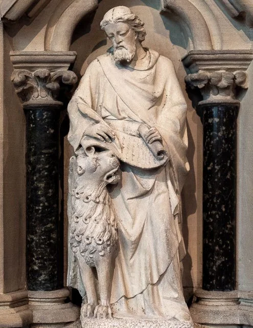 Statue of a bearded man with long hair, dressed in robes, holding a book and a staff, standing beside a lion.