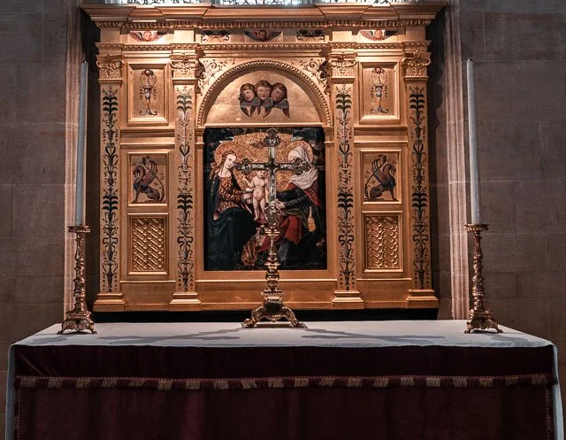Religious altar with gold-colored wooden frame, featuring a painting of Jesus on the cross with two women at his sides, and small paintings of saints or angels above. Two tall lit candles are on each side of the altar on a table covered with a white 
