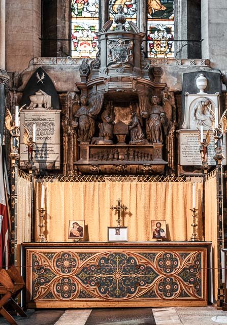 An ornate altar inside a church with religious statues, candles, framed images, and a cross, set against a backdrop of stained glass windows.