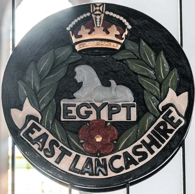 Emblem of Egypt East Lancashire mounted on a black circular background with a crown at the top, a horse in the center, green laurel branches surrounding, and a red flower at the bottom.