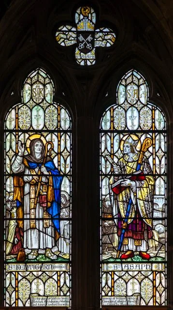 Colorful stained glass windows depicting two saints, Saint Keyne and Saint Asaph, inside a church.
