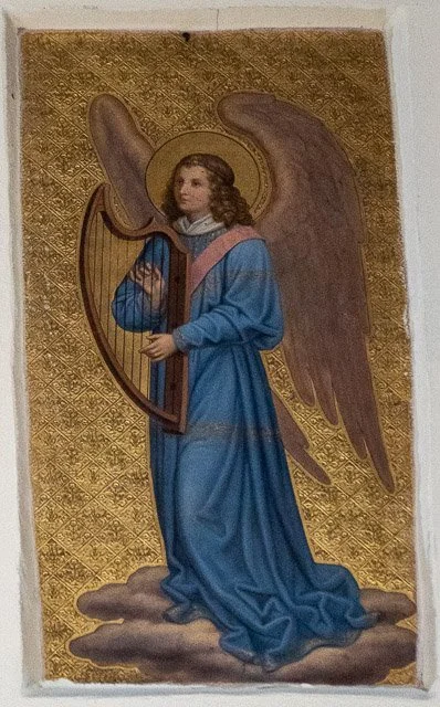 Religious painting of an angel with large wings, holding a harp, dressed in a blue robe, standing on a cloud with a golden patterned background.
