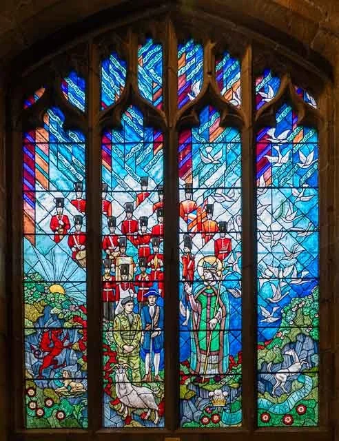 Colorful stained glass window with military figures, soldiers, and animals including a dog and horses, set in a church or cathedral.