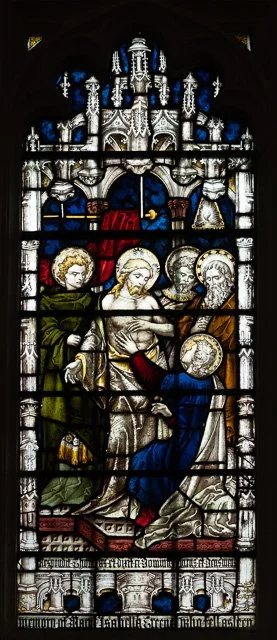 A stained glass window depicting a religious scene with four haloed figures, one kneeling and others standing around, in front of a church background.