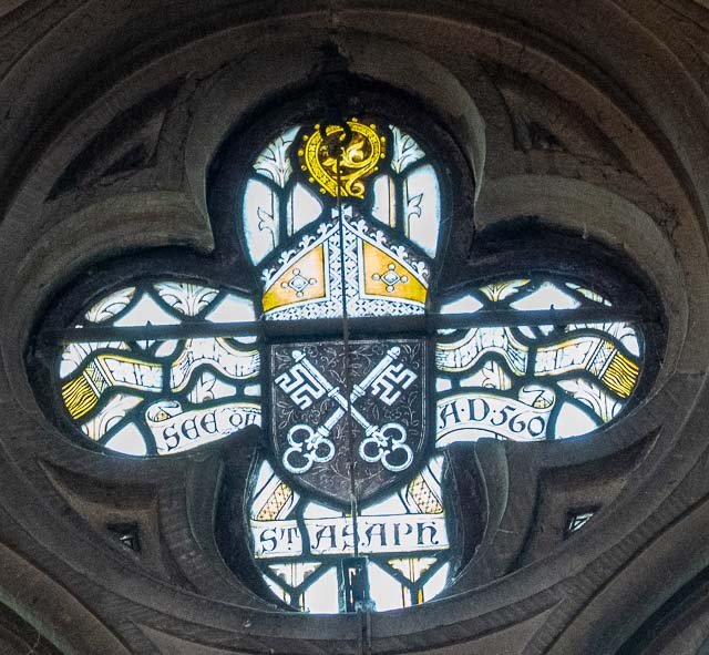 A stained glass clock with three black clock hands, decorated with symbols and Latin text, inside a stone window frame.
