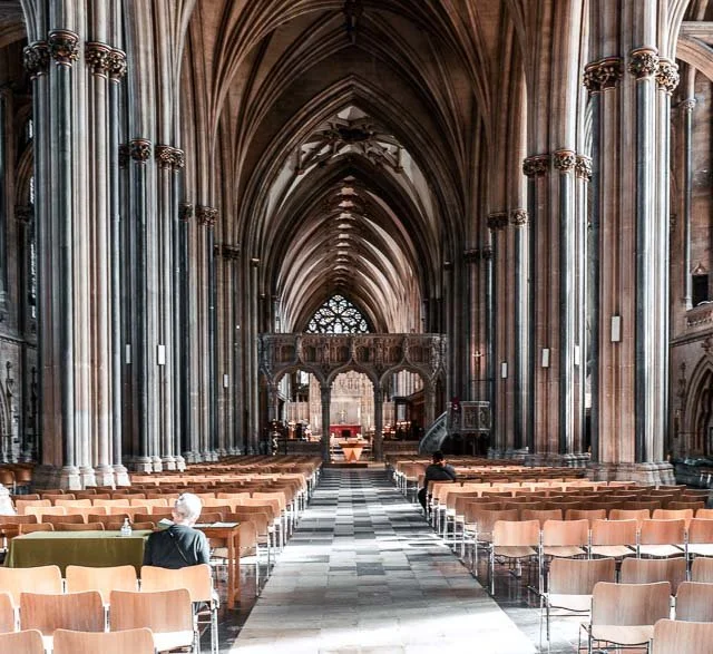 Interior of a large, ornate Gothic cathedral with high vaulted ceilings, tall columns, and rows of seating, with a few people seated.