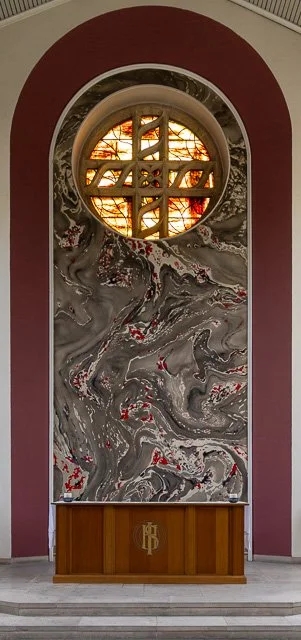 Interior of a church or chapel with a large circular stained glass window featuring a cross design, marble wall with swirling patterns, and a wooden altar at the bottom with a monogram.