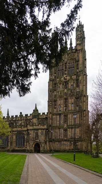 A historic stone church with a tall clock tower, surrounded by a park with trees and a brick pathway.