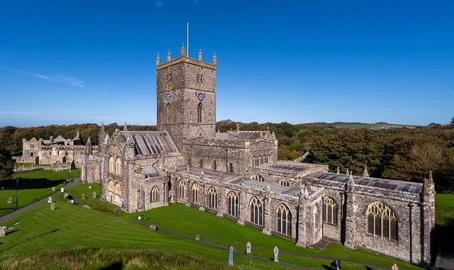 A historic stone church with a tall square bell tower, situated on a lush green hillside under a clear blue sky.