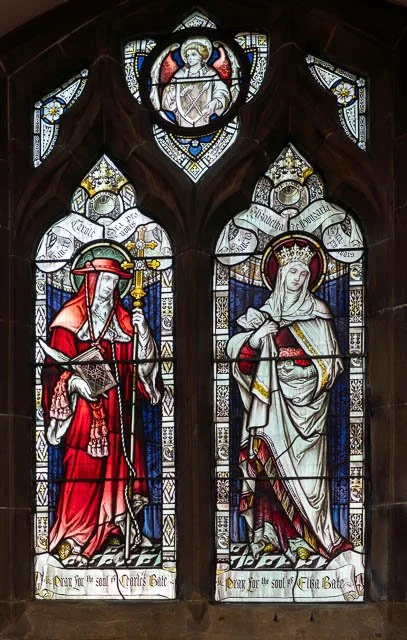 Stained glass window depicting a knight in armor and a crowned woman in white robe with cross staff, within gothic arches.