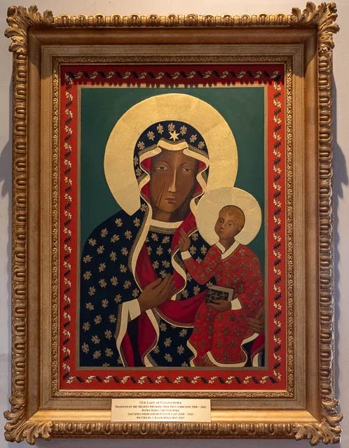 Religious icon painting of the Virgin Mary holding baby Jesus, both with golden halos, set against a green background with a red patterned border, in an ornate gold frame.