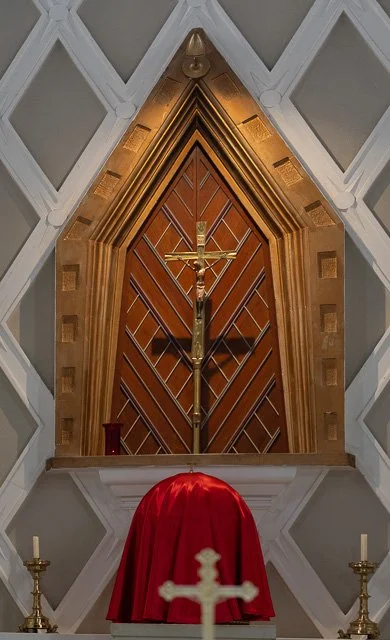A crucifix with Jesus on the cross, covered with a red cloth, flanked by two candles on gold stands in a church altar with a wooden backdrop and geometric wall design.