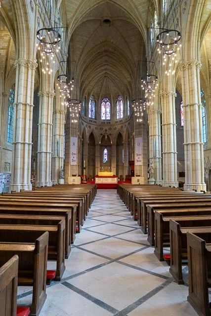 Interior of a church with high arched ceilings, wooden pews, hanging chandeliers, and stained glass windows.