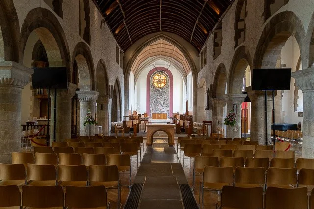 Interior of a church or chapel with wooden chairs arranged in rows facing an altar, stone arches lining the sides, and a high arched wooden ceiling. There are two large black speakers and some flowers near the columns. The back of the church features