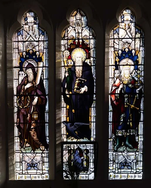 Three stained glass windows in a church, depicting religious figures with halos, detailed patterns, and decorative elements.