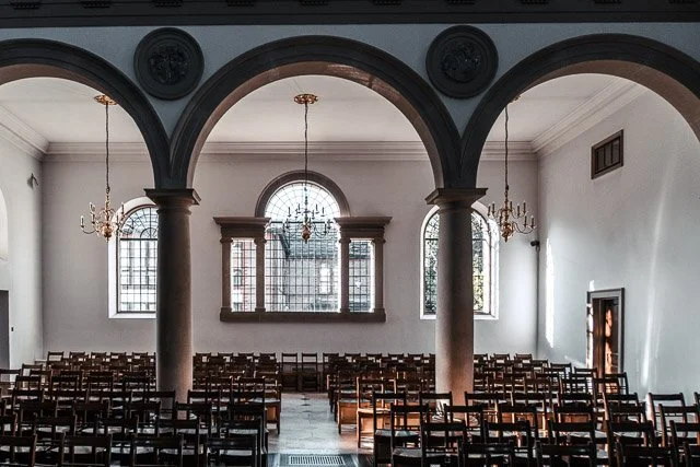 Interior of an empty church with high arched windows, columns, chandeliers, and rows of wooden chairs