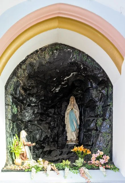 Religious shrine with a statue of the Virgin Mary in a rocky alcove, decorated with flowers and plants.