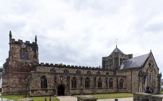 A large historic stone church with gothic windows, a tower with a clock, and a smaller tower with a weather vane, set against a cloudy sky.