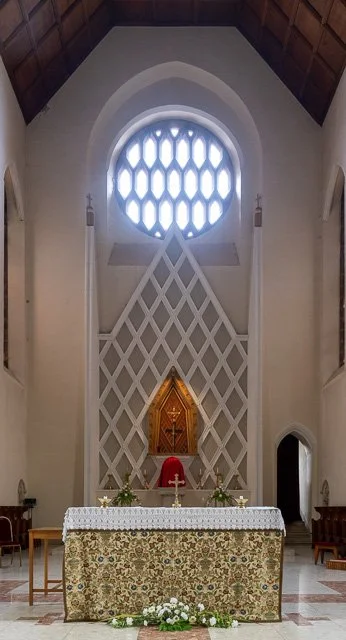 Inside a church with an altar covered by a decorated cloth, a wooden crucifix, and a red cloth, window with a honeycomb pattern of light, and tall side windows.