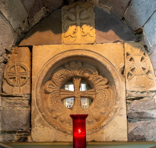 Stone wall with carved Christian crosses and symbols, including a circular cross and a chalice with a candle in front.