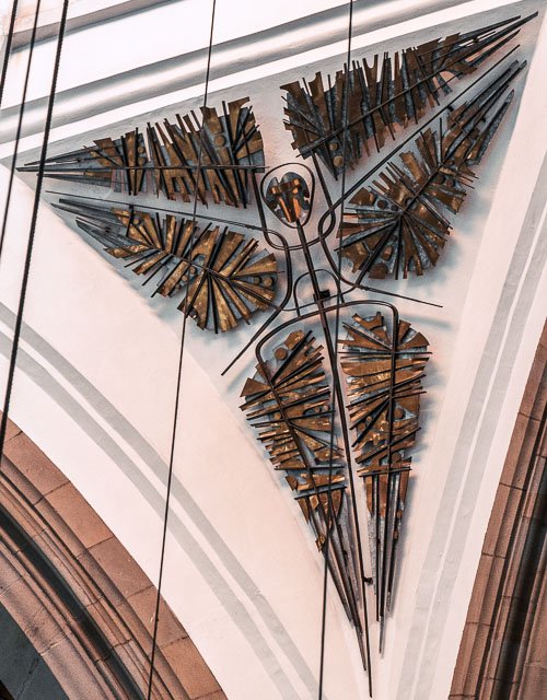 Artistic clock with a triangular face made of wooden sticks and metal hands mounted on a white wall.